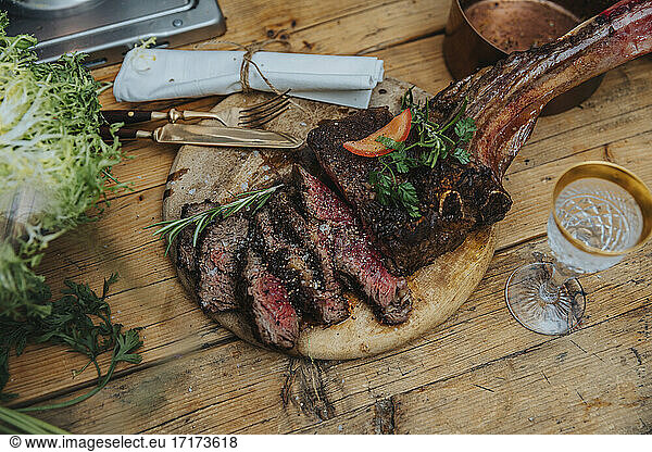 Tomahawk steak garnished with vegetables on wooden plate by drink in kitchen