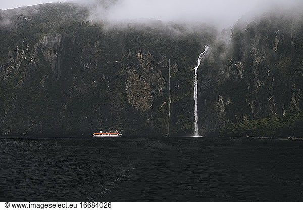Tourist ferry approaching waterfall at Milford Sound during foggy day.