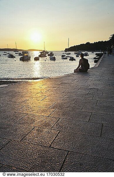 Tourist sitzt und beobachtet den Sonnenuntergang im Hafen der Stadt Hvar  Insel Hvar  Dalmatien  Kroatien. Dies ist ein Foto eines Touristen  der den Sonnenuntergang im Hafen der Stadt Hvar  Insel Hvar  Dalmatien  Kroatien  beobachtet. Es gibt viele Orte  an denen man einen Sonnenuntergang in Hvar beobachten kann  und nur wenige sind besser als der alte Hafen.
