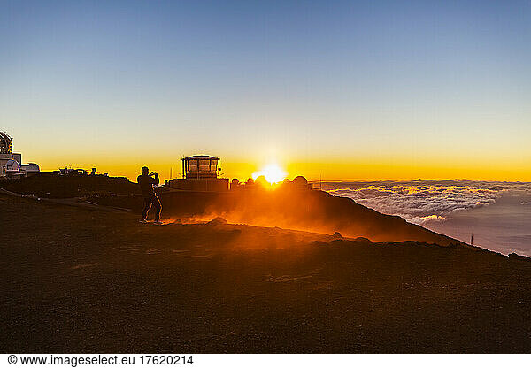 Tourist takes a photograph from the Haleakala Observatory above the clouds with a golden sunset and a view of the ocean below; Maui  Hawaii  United States of America