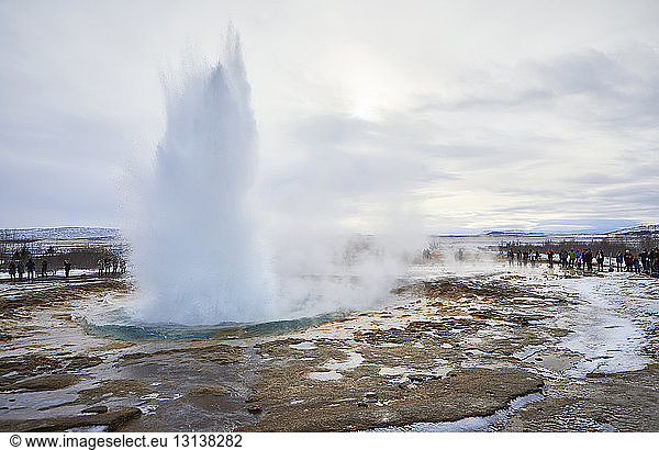 Tourists looking at water splashing from geyser against cloudy sky during winter