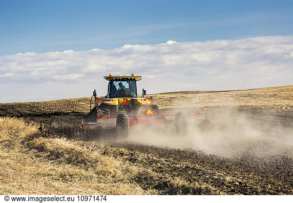 Tractor tilling a hill side field with clouds and blue sky; Alberta Canada