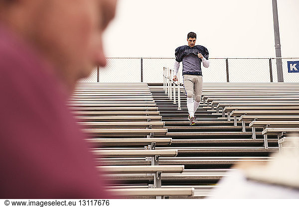 Trainer hält Klemmbrett  während ein American-Football-Spieler im Stadion trainiert