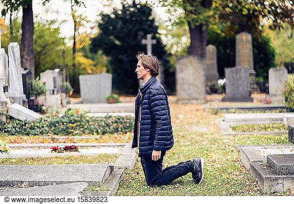 Trauernder junger Mann kniend vor einem Grab auf einem Friedhof