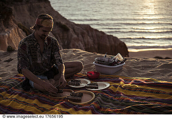 traveling man prepares picnic dinner by ocean as sunsets