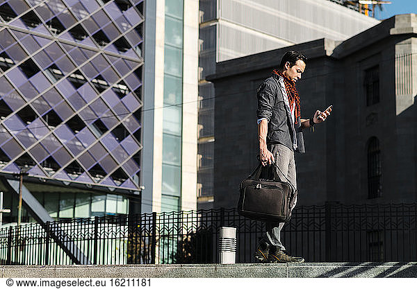 Trendy Mann mit Handtasche mit Smartphone beim Spaziergang in der Stadt