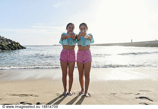 Twin sisters in matching outfits holding squirt guns on beach at sunny day