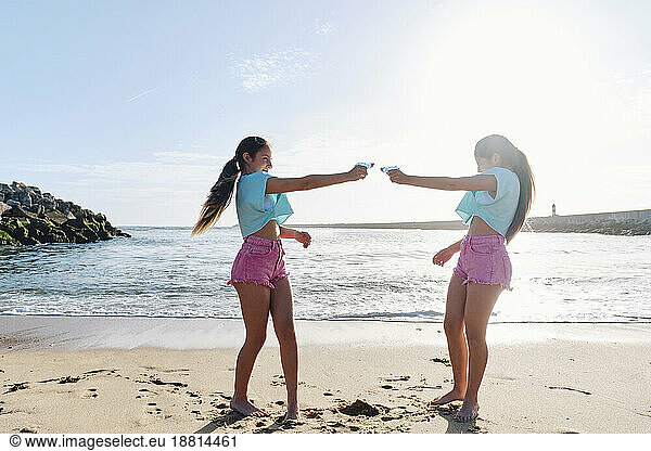 Twin sisters playing with squirt guns on beach at sunny day