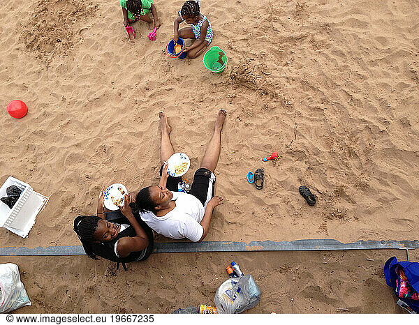 Two Black Women & 2 Girls on the beach eating and playing