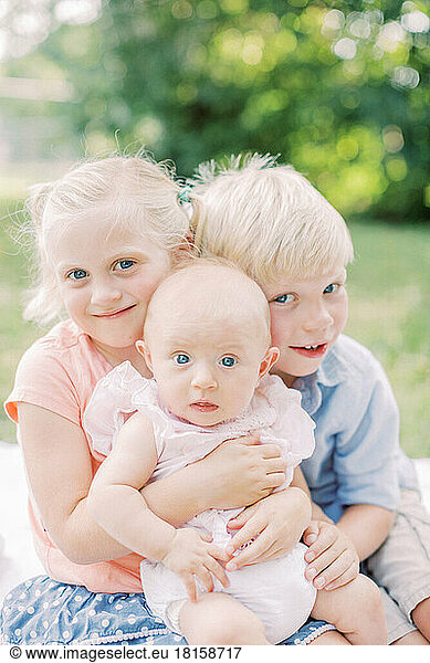 Two blonde toddlers hug their blue-eyed baby sister in park.