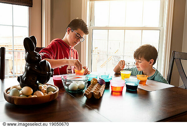 Two boys coloring Easter eggs with containers of dye at wooden table.