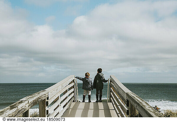 Two boys on boardwalk leading to beach against ocean and cloudy sky