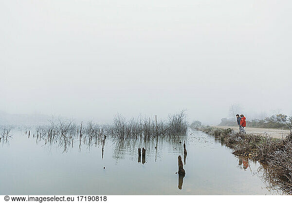 Two brothers standing beside lake in winter against foggy overcast sky