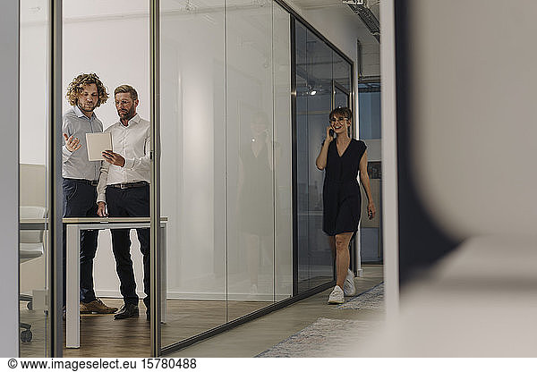 Two businessmen using tablet in office with businesswoman on the phone passing by