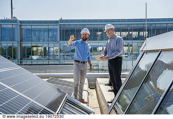 Two businessmen wearing hardhats having a meeting on the roof of a company building with solar panels