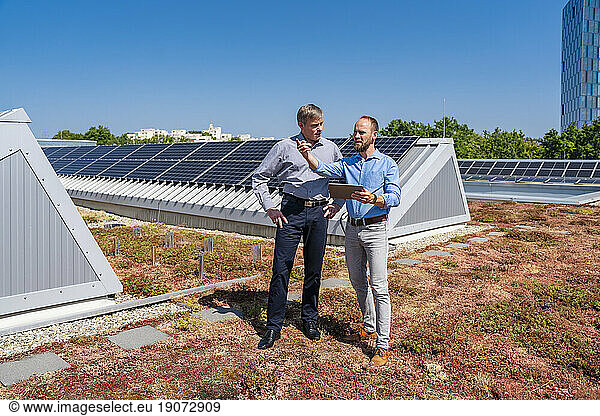 Two businessmen with tablet PC having a meeting on the roof of a company building with solar panels
