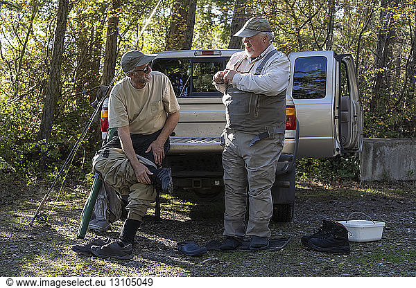 Two Caucasian male fly fishermen enjoy some conversation around a pickup truck while putting on their waders to go fly fishing.