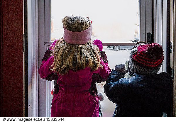 Two children dressed in winter clothing look out a door at snow.