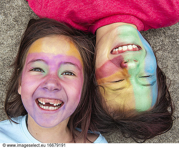 Two children lay down cheek-to-cheek with colorful face paint laughing