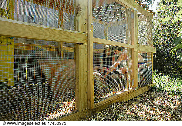 two children sit together inside enclosed run playing with chickens