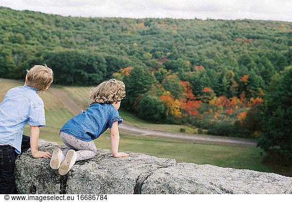 Two children sitting on a rock wall while looking at fall foliage