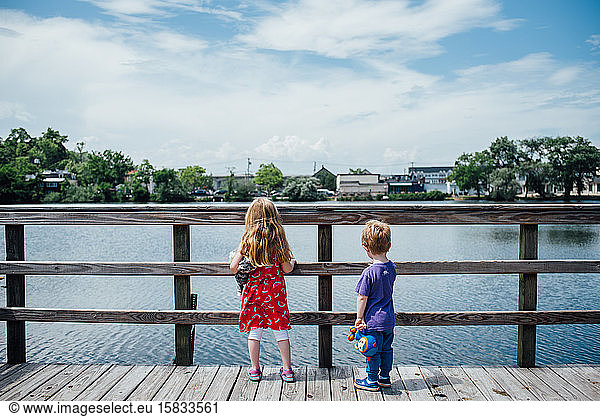 Two children stand at a pier and look out at a pond.