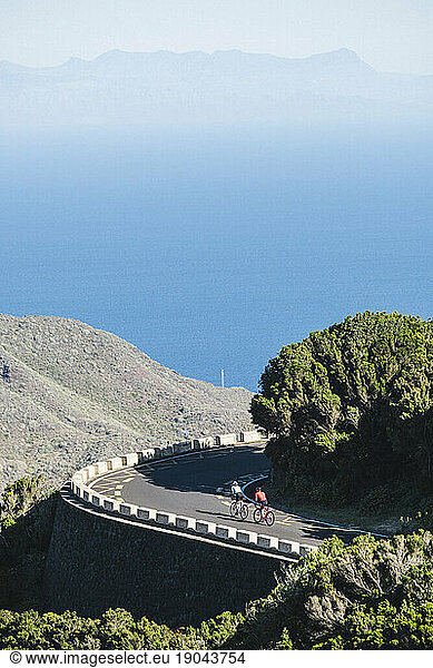 Two cyclists pulled back view on winding road with ocean in background