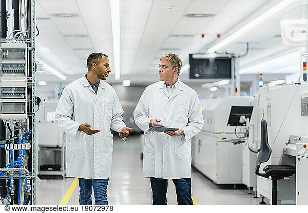 Two electrical technicians walking through factory workshop discussing over digital tablet