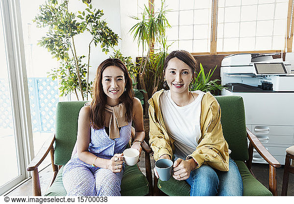 Two female Japanese professionals sitting in a co-working space  smiling at camera.