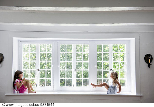 Two girls playing  sitting on a ledge by a large window.