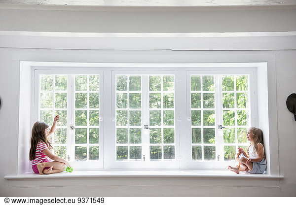Two girls playing  sitting on a ledge by a large window.