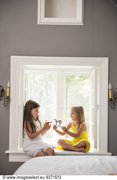 Two girls playing together  sitting on a window seat indoors.