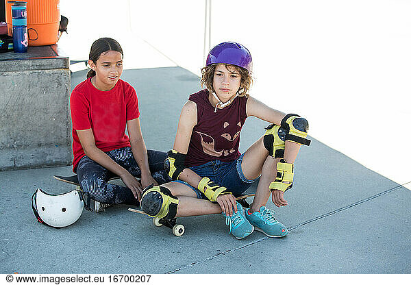 Two girls sit in the shade with their skateboards at the skatepark