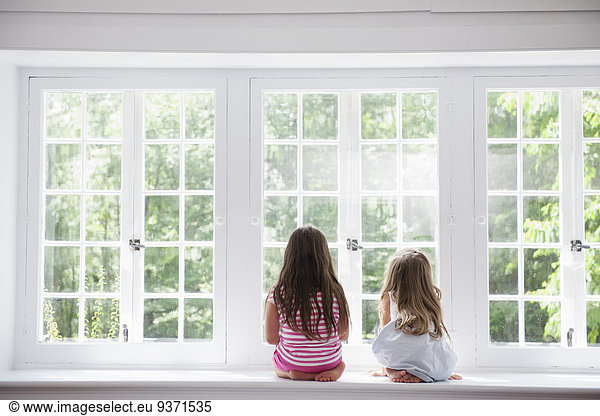 Two girls sitting side by side by a large window.