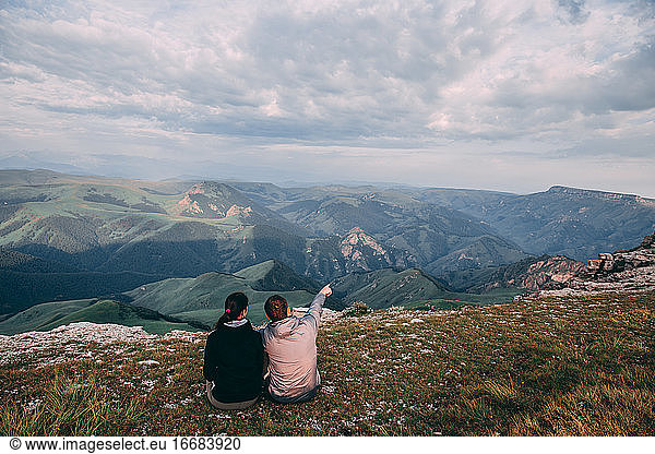 Two guys are sitting and looking into the distance and the mountains  discussing something and showing somewhere far into the distance with his hand.