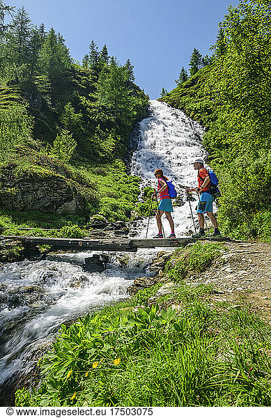 Two hikers crossing bridge over small waterfall in Hohe Tauern National Park