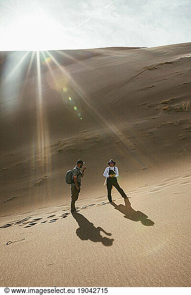 two hikers in sand dunes stop and smile under sun flare