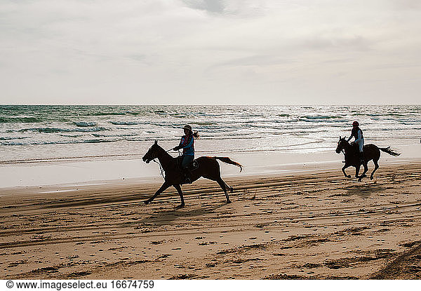 Two jockeys racing Andalusian horses backlit down beach