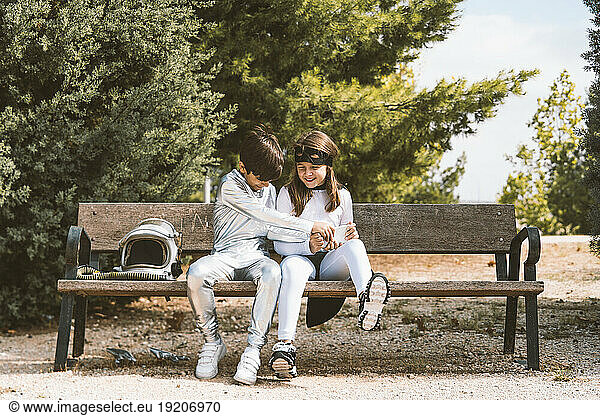 Two kids in astronaut and superhero costumes with mobile phone on park bench