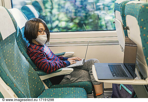 Two laptops and boy with mask uses computer while traveling by train