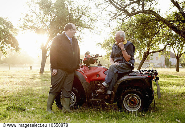 Two men  a farmer and a man with a clipboard  by a quadbike in an orchard.