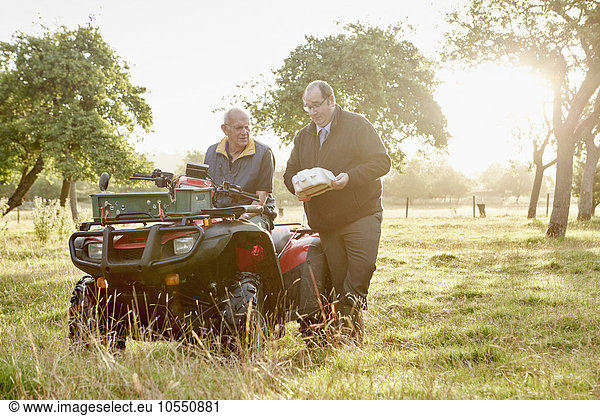 Two men  a farmer and a man with a clipboard  by a quadbike in an orchard.