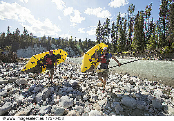 Two men carry inflatable rafts on a river adventure.
