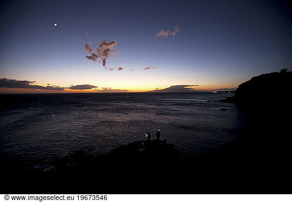 Two men fishing with headlamps as the sun sets over the ocean.