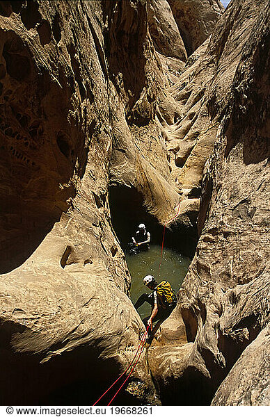 Two men going down desert canyon in wetsuites using ropes