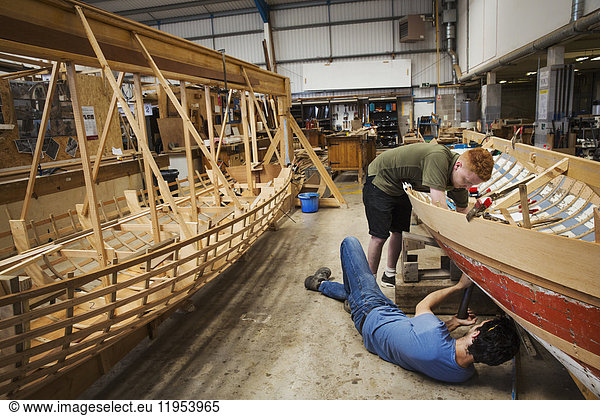 Two men in a boat-builder's workshop  working together on a wooden boat hull.