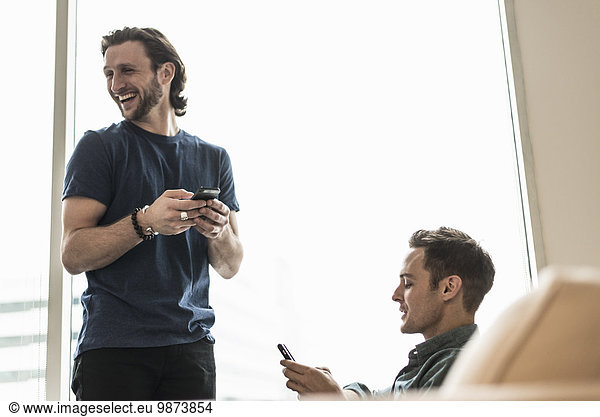 Two men in an office  checking their smart phones. One looking away laughing.