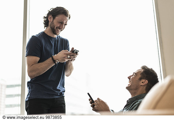 Two men in an office  checking their smart phones. One looking down laughing.