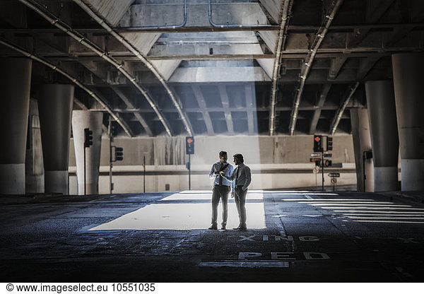 Two men in an urban underpass  with concrete and glass structures surrounding them  taking a selfie.