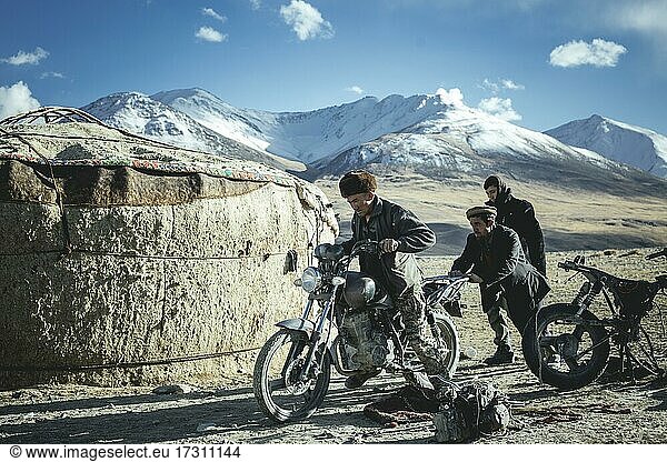 Two men push the motorcycle of a Kyrgyz nomad to get it running again  next to it spare parts and an engine of another motorcycle  in the background the snowy peaks of the Hindu Kush  Wakhan corridor  Badakhshan  Afghanistan  Asia